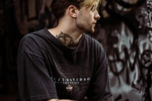 A tattooed man in a black T-shirt sits in a graffiti-covered urban environment, showcasing street style.