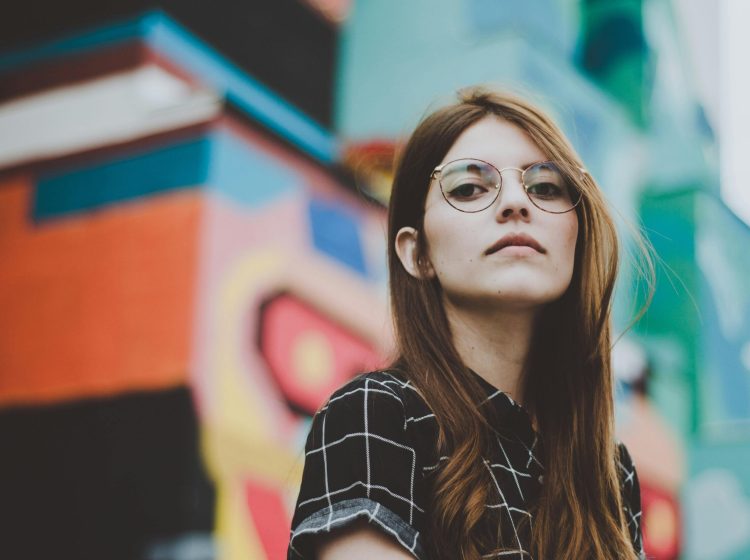 Portrait of a fashionable woman with glasses posing confidently outdoors in front of a vibrant mural.