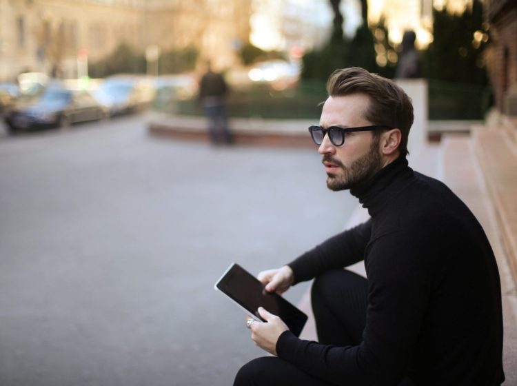 Side view of trendy serious bearded male in glasses and black casual clothes with notebook looking away and thinking while sitting on exterior stairs at entrance of modern building in downtown