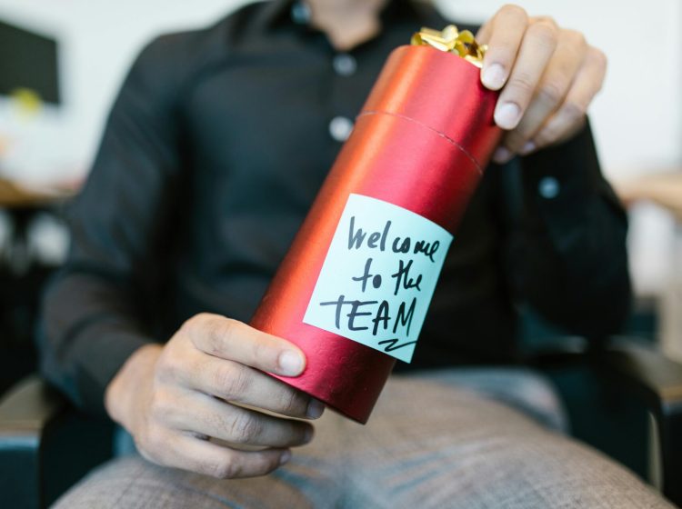 A red gift box held by a person with a welcome to the team note, ideal for new hire announcements.