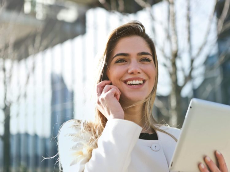 Confident businesswoman using her tablet and phone, smiling outdoors in sunlight.